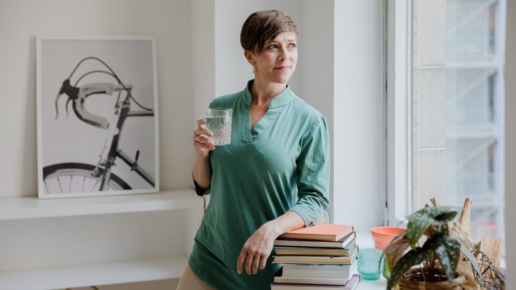Woman standing indoors with books and a glass of water near a window.